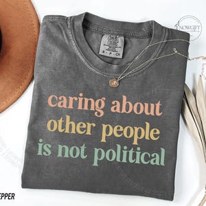 May include: A gray t-shirt with the text "caring about other people is not political" printed in colorful letters. The shirt has a distressed look and is made of a soft, comfortable fabric.
