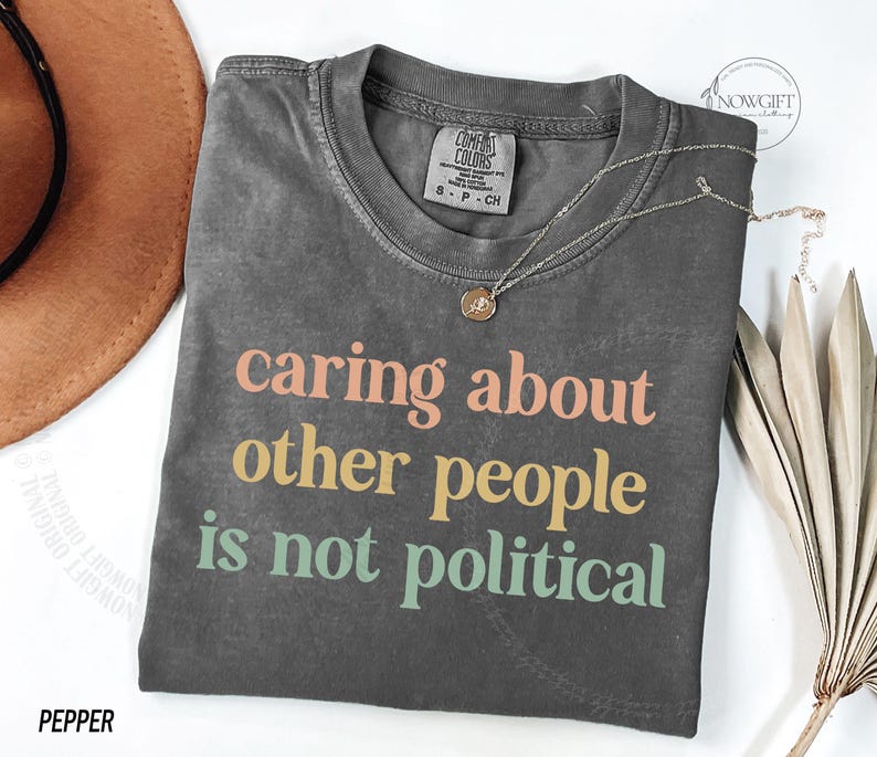May include: A gray t-shirt with the text "caring about other people is not political" printed in colorful letters. The shirt has a distressed look and is made of a soft, comfortable fabric.