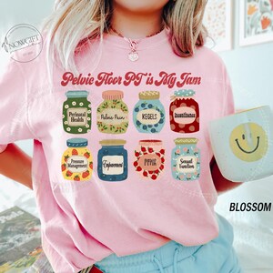 May include: Pink sweatshirt with the text "Pelvic Floor PT is My Jam" and illustrations of jam jars with labels like "Perinatal Health" and "Kegels." A mug with a smiley face is held in the image.