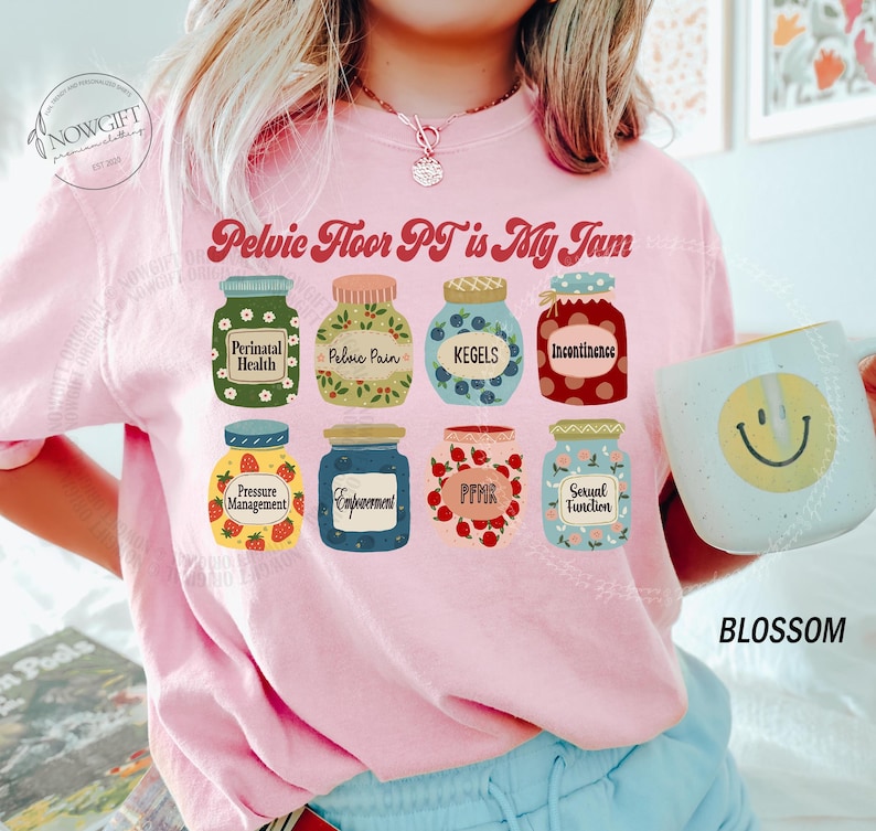 May include: Pink sweatshirt with the text "Pelvic Floor PT is My Jam" and illustrations of jam jars with labels like "Perinatal Health" and "Kegels." A mug with a smiley face is held in the image.