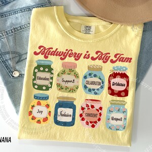 May include: A yellow t-shirt with the text "Midwifery is My Jam" in red script. The shirt features illustrations of jars with labels like "Education," "Support," and "Collaboration." The shirt is made of a soft, comfortable material.