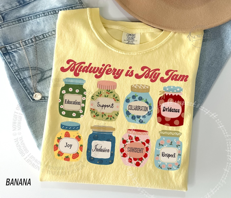 May include: A yellow t-shirt with the text "Midwifery is My Jam" in red script. The shirt features illustrations of jars with labels like "Education," "Support," and "Collaboration." The shirt is made of a soft, comfortable material.