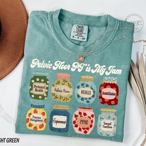 May include: Light green t-shirt with the text "Pelvic Floor PT is My Jam" and illustrations of jam jars labeled with terms like "Perinatal Health" and "Kegels." A silver necklace with a pendant is draped on the shirt.