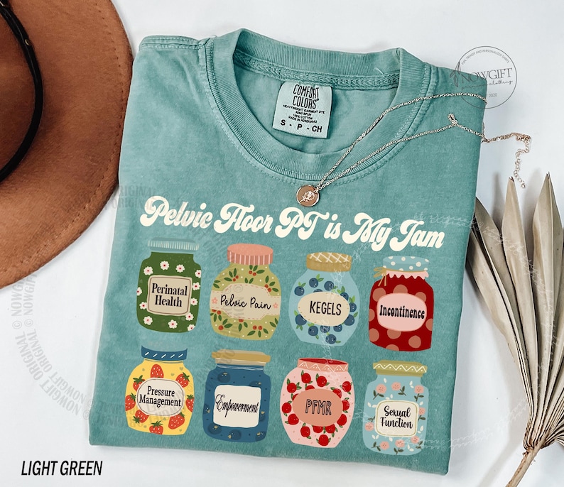 May include: Light green t-shirt with the text "Pelvic Floor PT is My Jam" and illustrations of jam jars labeled with terms like "Perinatal Health" and "Kegels." A silver necklace with a pendant is draped on the shirt.
