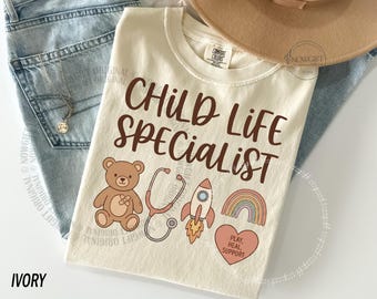 Op de afbeelding: Ivoor kleurig t-shirt met de tekst "CHILD LIFE SPECIALIST" in bruin script. Het shirt toont illustraties van een teddybeer, stethoscoop, raket, regenboog en een hart met de woorden "PLAY, HEAL, SUPPORT". Het shirt is op een licht oppervlak gevouwen.