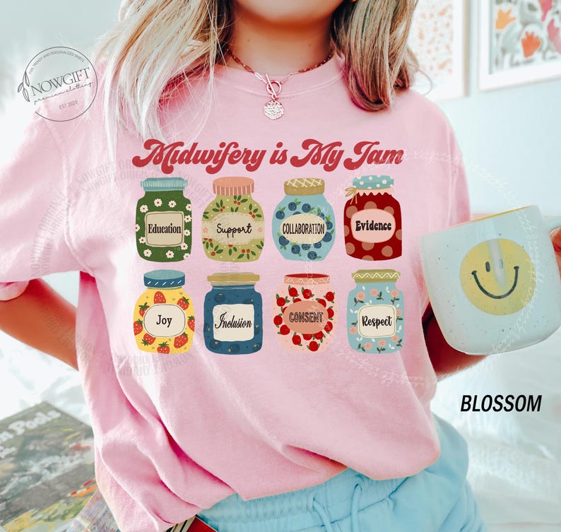 May include: Pink sweatshirt with the text "Midwifery is My Jam" and illustrations of jars labeled with words like "Education," "Support," and "Joy." A mug with a smiley face is held in the image.