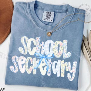 May include: A blue Comfort Colors t-shirt with the words "School Secretary" in a floral and gingham pattern. A light blue bow is centered below the word "School". A gold necklace with a pendant is on the shirt.