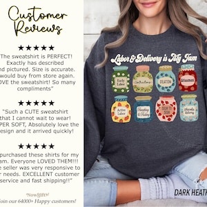 May include: A dark heather grey sweatshirt with the text "Labor & Delivery is My Jam" printed on the front. The text is surrounded by nine illustrated jars, each representing a different stage of labor. The jars are decorated with flowers, berries, and other details.