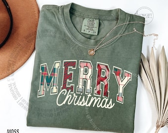 Könnte beinhalten: Ein moosgrünes T-Shirt mit den Worten "MERRY Christmas" in einem Karomuster. Das Shirt hat einen Rundhalsausschnitt und kurze Ärmel. Eine goldene Halskette mit Anhänger liegt über dem Shirt. Ein brauner Hut befindet sich in der oberen linken Ecke.