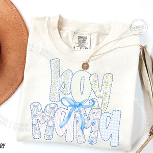 Puede incluir: Camiseta Comfort Colors color marfil con la frase "Boy Mama" en un dise&ntilde;o de patchwork. Las letras est&aacute;n rellenas de estampados florales y cuadros vichy, y un lazo azul adorna la palabra "MAMA". Tambi&eacute;n se ve un collar dorado y un sombrero.