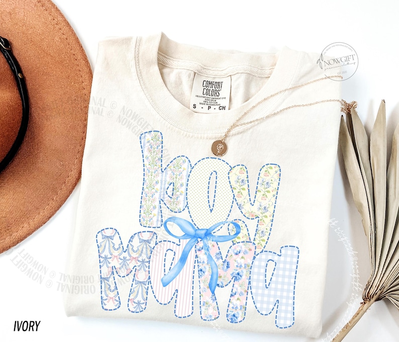 Puede incluir: Camiseta Comfort Colors color marfil con la frase "Boy Mama" en un dise&ntilde;o de patchwork. Las letras est&aacute;n rellenas de estampados florales y cuadros vichy, y un lazo azul adorna la palabra "MAMA". Tambi&eacute;n se ve un collar dorado y un sombrero.