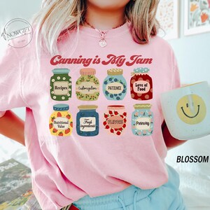 Peut inclure: T-shirt rose avec le texte "Canning is My Jam" et une rang&eacute;e de bocaux illustr&eacute;s avec diff&eacute;rentes &eacute;tiquettes, notamment "Recettes", "Personnalisation", "Patience", "Amour de la nourriture", "Valeur nutritionnelle", "Ingr&eacute;dients frais", "Saveurs" et "Conservation".