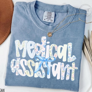 May include: Blue jean Comfort Colors t-shirt with the words "Medical Assistant" in a patchwork design. The letters are white with floral and gingham patterns. A gold necklace and a brown hat are also visible.