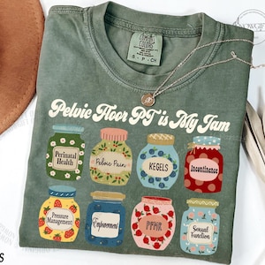 May include: Moss green t-shirt with the text "Pelvic Floor PT is My Jam." The shirt features illustrations of jars labeled with terms like "Perinatal Health," "Pelvic Pain," and "Kegels." A gold necklace is draped across the shirt.