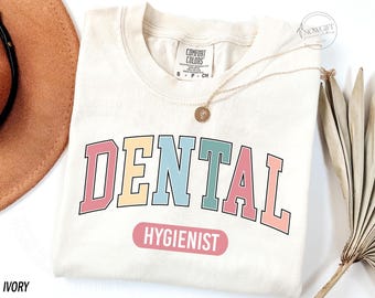 May include: Ivory coloured t-shirt with the text "DENTAL HYGIENIST" in rainbow colours. The text is in a varsity style font.