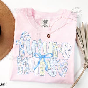 May include: A light pink t-shirt with the words "FUTURE NURSE" in a patchwork design with blue floral and gingham patterns. A blue bow is centered below the word "FUTURE". A gold necklace with a small pendant is draped on the shirt.