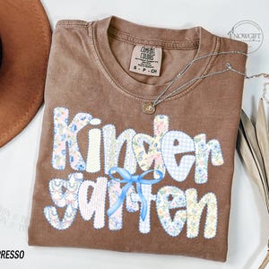 Pu&ograve; includere: T-shirt Comfort Colors color espresso con la scritta "Kindergarten" in un design patchwork. Le lettere presentano motivi floreali e a quadri, con un fiocco azzurro. Una collana d'argento con un ciondolo &egrave; drappeggiata sulla maglietta.