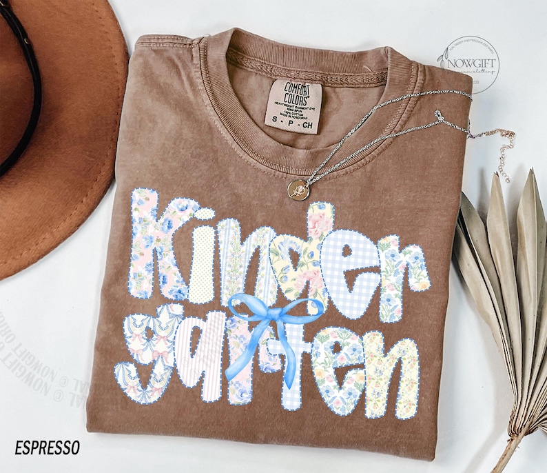 Pu&ograve; includere: T-shirt Comfort Colors color espresso con la scritta "Kindergarten" in un design patchwork. Le lettere presentano motivi floreali e a quadri, con un fiocco azzurro. Una collana d'argento con un ciondolo &egrave; drappeggiata sulla maglietta.