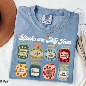 May include: A blue t-shirt with the slogan "Books are My Jam" and illustrated jam jars. Each jar has a different label, including "Endless TBR" and "Bookmarks". A gold necklace is draped across the shirt.