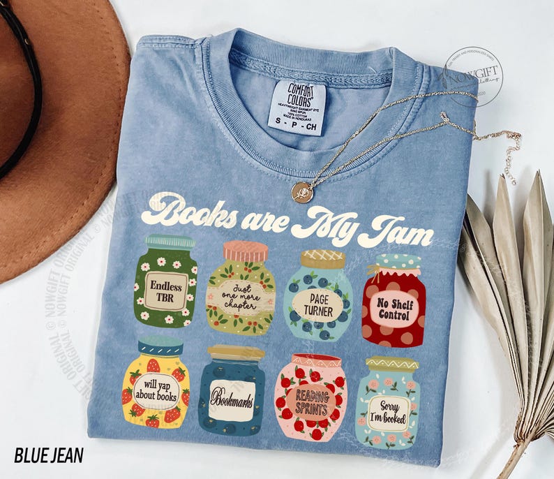 May include: A blue t-shirt with the slogan "Books are My Jam" and illustrated jam jars. Each jar has a different label, including "Endless TBR" and "Bookmarks". A gold necklace is draped across the shirt.
