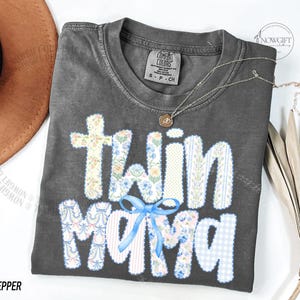 Puede incluir: Una camiseta gris Comfort Colors con las palabras "Twin Mama" en un dise&ntilde;o de patchwork. Las letras est&aacute;n decoradas con patrones florales y de cuadros, y un lazo azul. Tambi&eacute;n se ve un collar plateado con un colgante.
