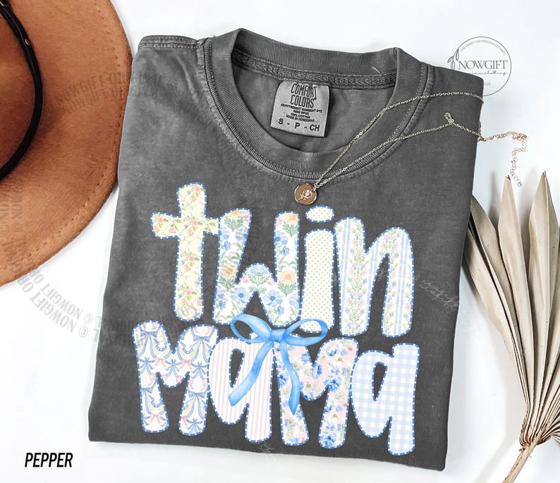 Puede incluir: Una camiseta gris Comfort Colors con las palabras "Twin Mama" en un dise&ntilde;o de patchwork. Las letras est&aacute;n decoradas con patrones florales y de cuadros, y un lazo azul. Tambi&eacute;n se ve un collar plateado con un colgante.