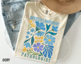 Op de afbeelding: Een ivoorkleurig t-shirt met de tekst "SPEECH-LANGUAGE PATHOLOGIST" in blauwe letters. Het ontwerp bevat een kleurrijk bloemstuk met blauwe, gele en paarse bloemen en groene bladeren gedrukt op de voorzijde van het kledingstuk.