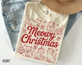 Op de afbeelding: Ivoorkleurig T-shirt met een rode "Meowy Christmas" opdruk met katten, kerstbomen en cadeaus. Het shirt ligt op een blauwe spijkerbroek en een beige hoed. Een feestelijk item.