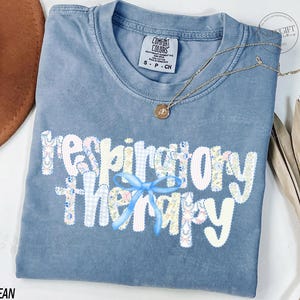 Pu&ograve; includere: Una maglietta blu jeans Comfort Colors con le parole "respiratory therapy" in un design patchwork. Le lettere sono bianche con motivi floreali e gingham. Un fiocco azzurro &egrave; centrato sotto la parola "therapy". Una collana d'oro e una pianta secca sono anche nell'immagine.