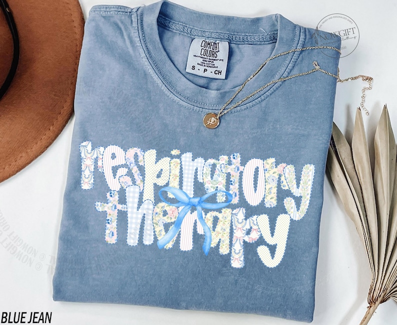Pu&ograve; includere: Una maglietta blu jeans Comfort Colors con le parole "respiratory therapy" in un design patchwork. Le lettere sono bianche con motivi floreali e gingham. Un fiocco azzurro &egrave; centrato sotto la parola "therapy". Una collana d'oro e una pianta secca sono anche nell'immagine.