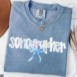 Könnte beinhalten: Ein blaues Comfort Colors T-Shirt mit dem Wort "Songwriter" in einem floralen Design und einer blauen Schleife. Eine goldene Halskette mit Anhänger befindet sich auf dem Shirt. Das Shirt liegt auf einer weißen Oberfläche.