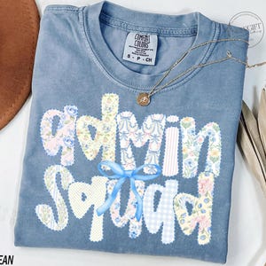 May include: A blue jean Comfort Colors t-shirt with the words "Admin Squad" in a patchwork design. The letters are made of floral and patterned fabrics. A blue ribbon is tied below the word "Squad". A gold necklace with a pendant is draped on the shirt.