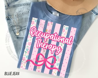Op de afbeelding: Een blauw T-shirt met korte mouwen en een grafische print van verticale strepen in afwisselende roze tinten en bloemmotieven. De tekst Occupational Therapy staat in een vetgedrukt roze cursief lettertype boven een roze strik.