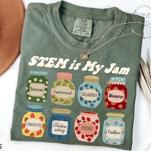 May include: A green t-shirt with the text "STEM is My Jam" in a retro font. The shirt features a design of colourful jars with words like "Innovate", "Experiment", "Collaborate", "Discover", "Communicate", "Problem Solving", "Design", and "Creative" on them.