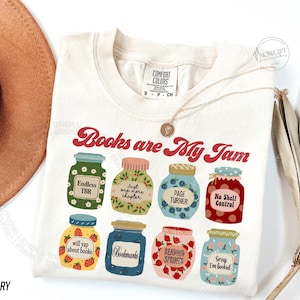 May include: Cream-coloured t-shirt featuring the text "Books are My Jam" in red script. The design includes illustrated jars with book-related labels such as "Endless TBR" and "Bookmarks". A gold necklace and a hat are also in the image.