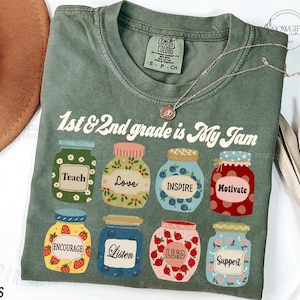 May include: A green t-shirt with the text "1st & 2nd grade is My Jam" in a retro font. The shirt features a design of nine jars with different words on them, including "Teach", "Love", "Inspire", "Motivate", "Encourage", "Listen", "Lead", and "Support".