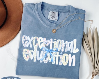 Op de afbeelding: Een kortmouwig t-shirt in spijkerblauw met een ronde hals. De voorkant toont de tekst "exceptional education" in een wit lettertype met een bloemen- en vichy-patroon. Een lichtblauwe lintstrik is over de letter "i" geplaatst.