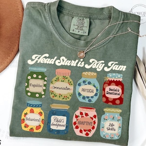 May include: Green t-shirt with the text "Head Start is My Jam" printed on it. The text is surrounded by illustrations of jars with different labels, including "Cognitive", "Communication", "Physical", "Social & Emotional", "Behavioral", "Child's Development", "Adaptive", and "Life Skills".