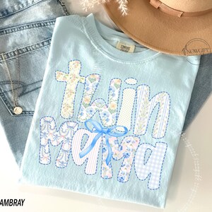 Puede incluir: Camiseta azul claro con la frase "Twin Mama" en letras de patchwork, con estampados florales y a rayas, y un lazo azul. La camiseta est&aacute; colocada sobre unos vaqueros azul claro, con un collar dorado visible. Un sombrero beige est&aacute; en el fondo.