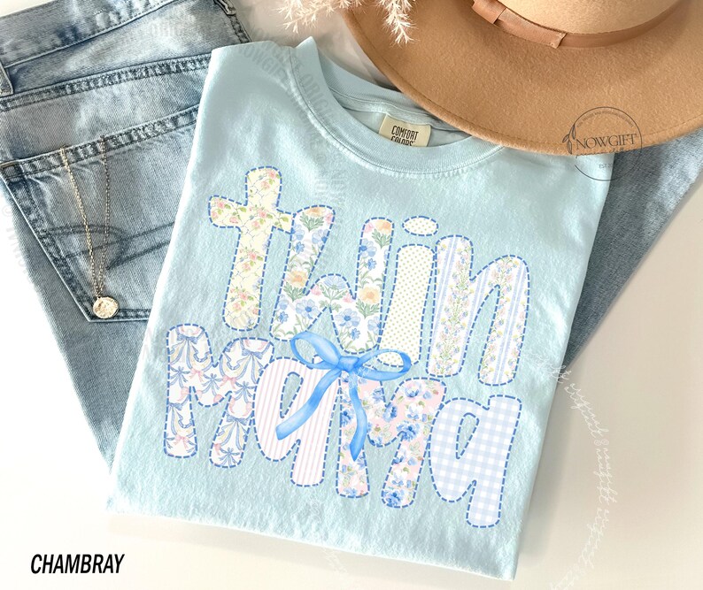Puede incluir: Camiseta azul claro con la frase "Twin Mama" en letras de patchwork, con estampados florales y a rayas, y un lazo azul. La camiseta est&aacute; colocada sobre unos vaqueros azul claro, con un collar dorado visible. Un sombrero beige est&aacute; en el fondo.