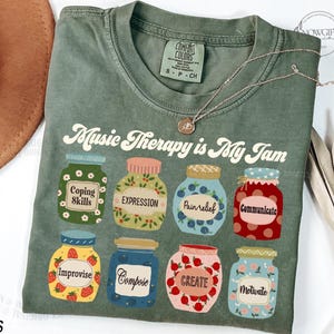 May include: A green t-shirt with the text "Music Therapy is My Jam" printed on it. The text is in a retro font. There are eight illustrated jars with different labels, such as "Coping Skills", "Expression", "Pain Relief", "Communicate", "Improvise", "Compose", "Create", and "Motivate".