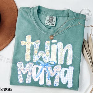 Puede incluir: Camiseta verde claro con la frase "Twin Mama" en un dise&ntilde;o de patchwork. Las letras est&aacute;n decoradas con estampados florales y un lazo azul. Un collar dorado con un colgante descansa sobre la camiseta.