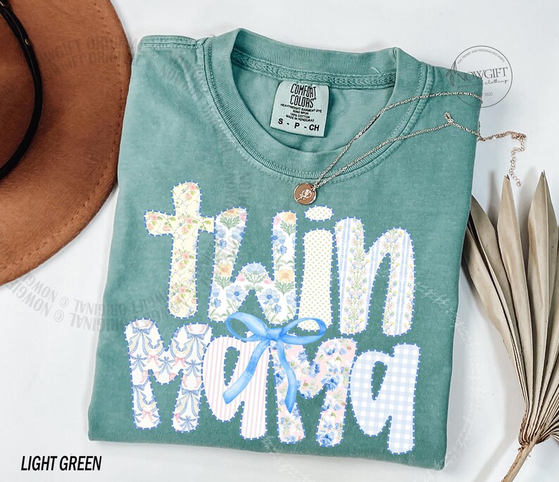 Puede incluir: Camiseta verde claro con la frase "Twin Mama" en un dise&ntilde;o de patchwork. Las letras est&aacute;n decoradas con estampados florales y un lazo azul. Un collar dorado con un colgante descansa sobre la camiseta.