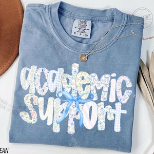 Peut inclure: Un t-shirt bleu jean Comfort Colors avec les mots "Academic Support" dans un motif patchwork. Les lettres sont décorées de motifs floraux et vichy, et un ruban bleu est noué en dessous. Un collier en or est drapé sur le t-shirt.