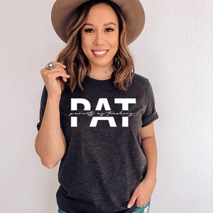 May include: A dark grey t-shirt with white text that reads "PAT parents as teachers". The person wearing the shirt is smiling and has brown hair. They are wearing a brown hat and blue jeans.