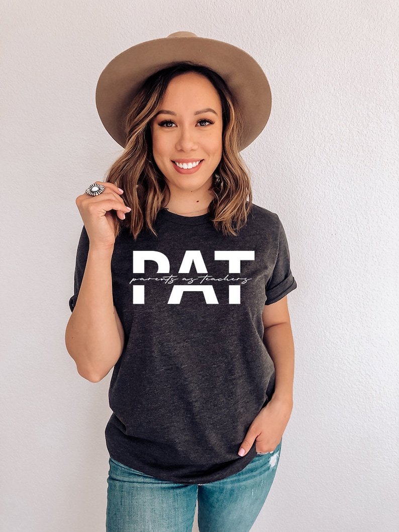 May include: A dark grey t-shirt with white text that reads "PAT parents as teachers". The person wearing the shirt is smiling and has brown hair. They are wearing a brown hat and blue jeans.