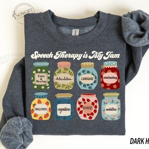 May include: Dark heather grey sweatshirt with the text "Speech Therapy is My Jam" in white. The design features nine colorful jars with speech therapy related words like "voice & hearing", "articulation", "language", "stuttering", "swallowing", "cognition", "social", and "modalities".