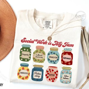 May include: Ivory t-shirt with the text "Social Work is My Jam" in red and white. The shirt features a design of nine jars with different labels, including "Empathy", "Cultural Awareness", "Organization", "Advocacy", "Communication", "Problem Solving", "Support", and "Active Listener".
