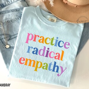 May include: A light blue t-shirt with the words "practice radical empathy" in colorful letters. The shirt is folded on top of a pair of light blue jeans and a tan hat. A gold necklace is visible.