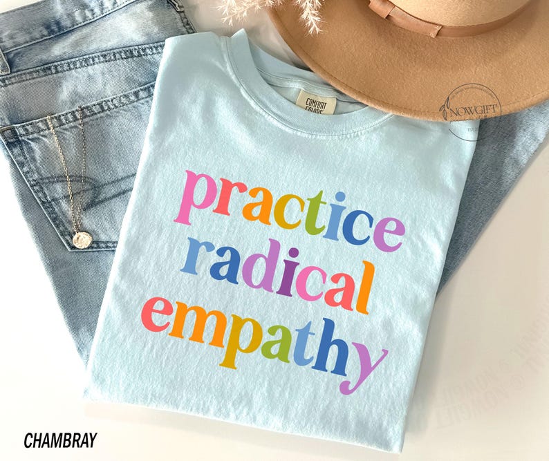 May include: A light blue t-shirt with the words "practice radical empathy" in colorful letters. The shirt is folded on top of a pair of light blue jeans and a tan hat. A gold necklace is visible.